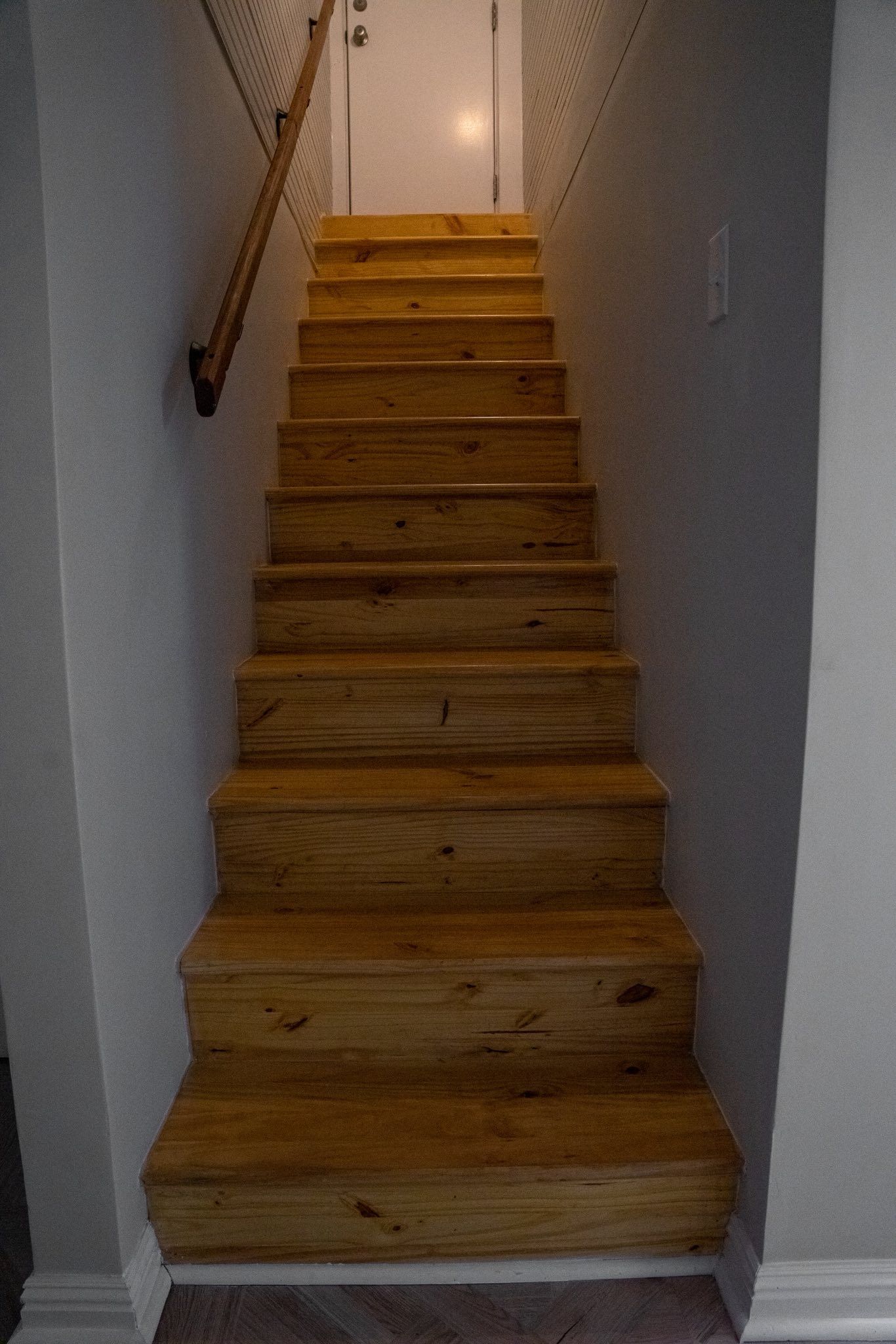 A straight, narrow wooden staircase leads upward between two gray walls to a closed white door.