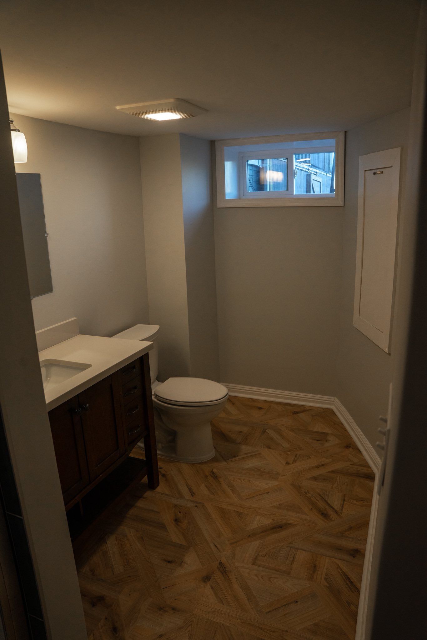 A bathroom with light gray walls, a wooden vanity, a white toilet, and brown patterned flooring under a recessed light.
