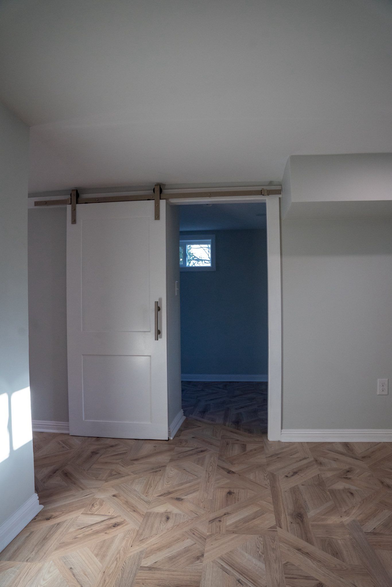 A white barn door installed on a metal track separating a room with herringbone wood flooring from a dark interior space.