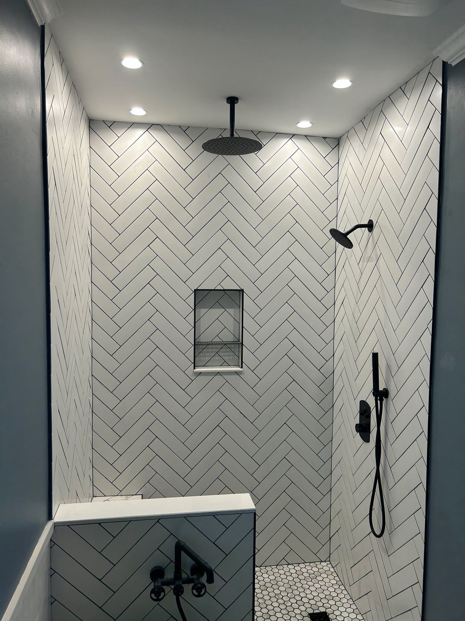 A shower interior with white herringbone tiles, a black rainfall showerhead, a handheld sprayer, and a built-in shelf.