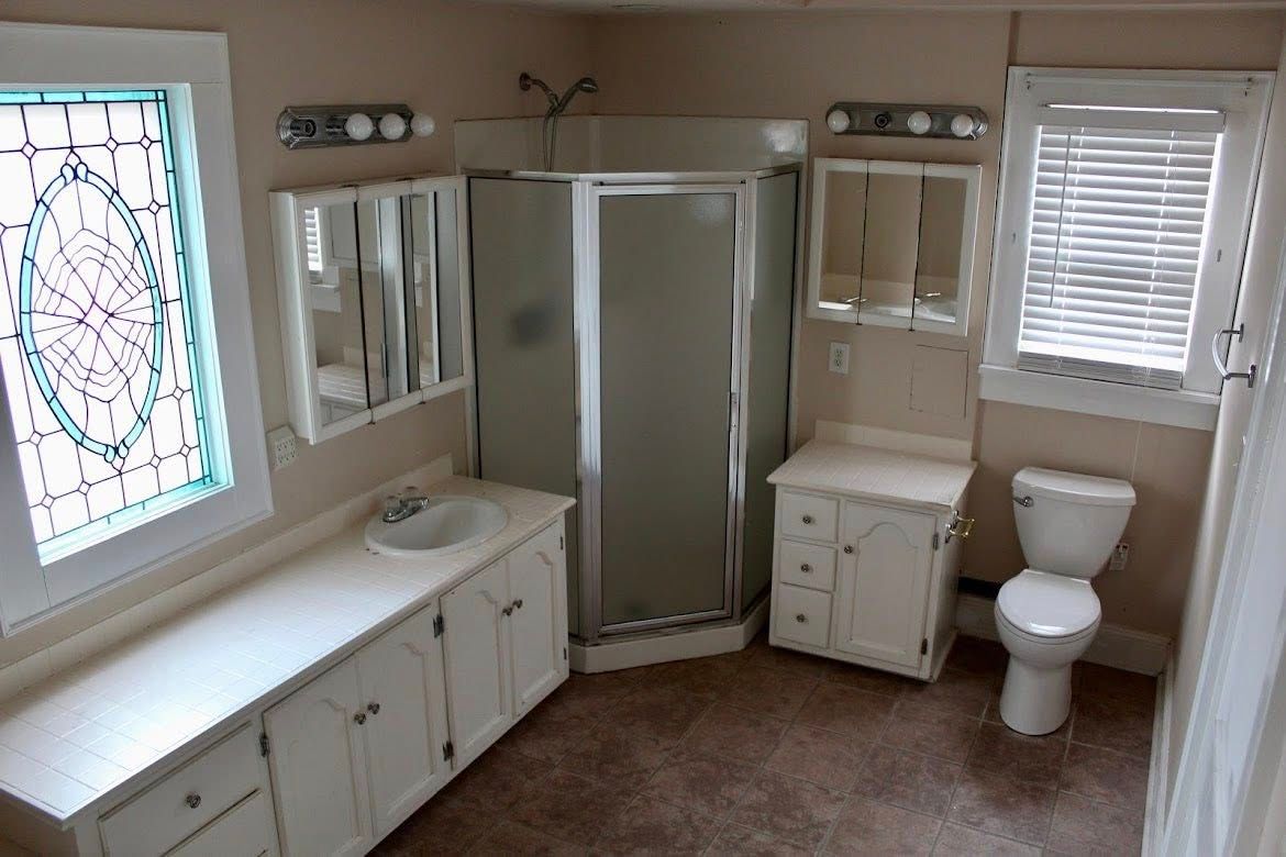 A bathroom featuring a white vanity with a sink, a corner shower, a second small vanity, and a toilet under a window.
