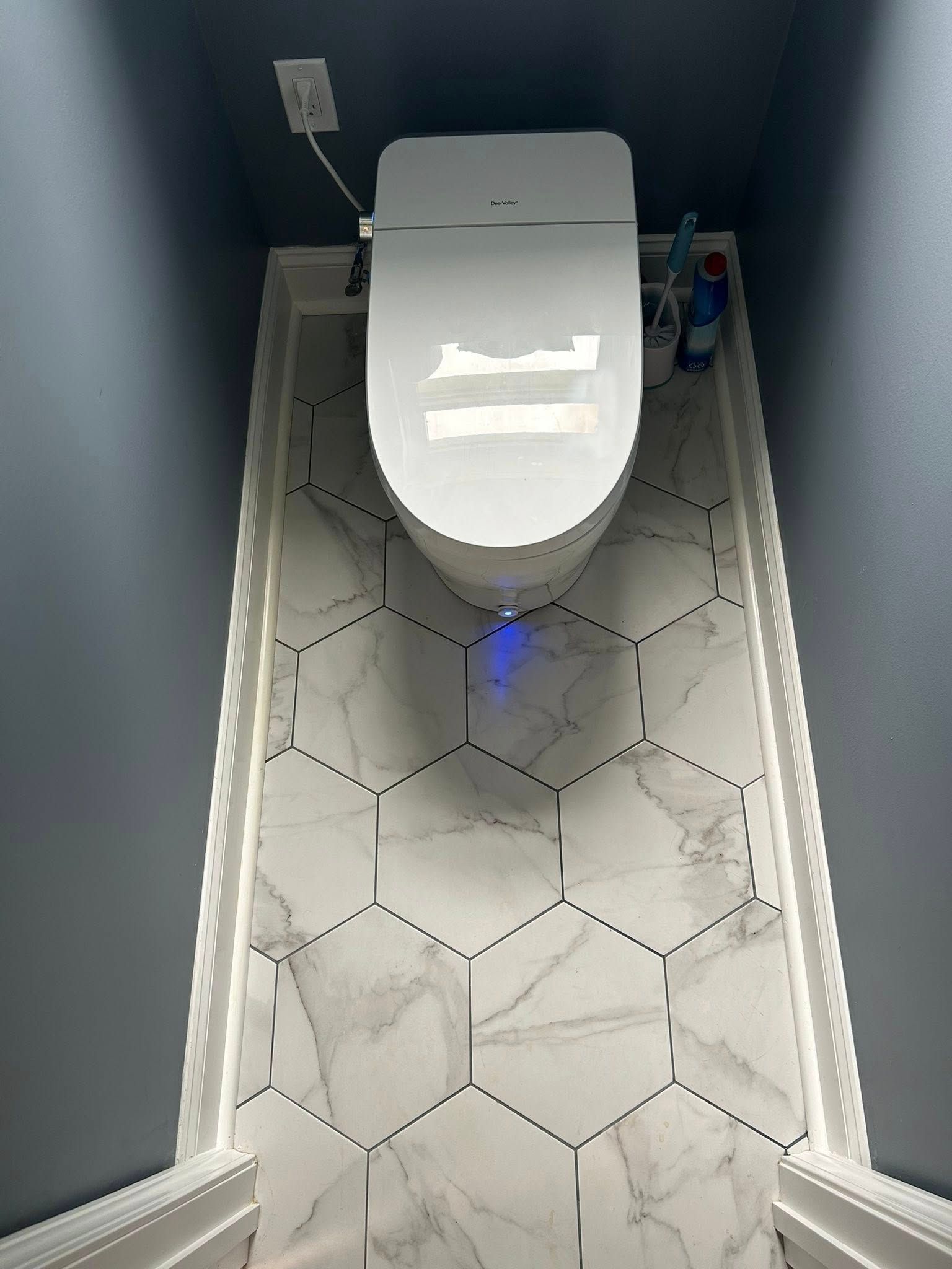 A smart toilet with a blue nightlight in a small room with dark gray walls and white hexagon marble floor tiles.