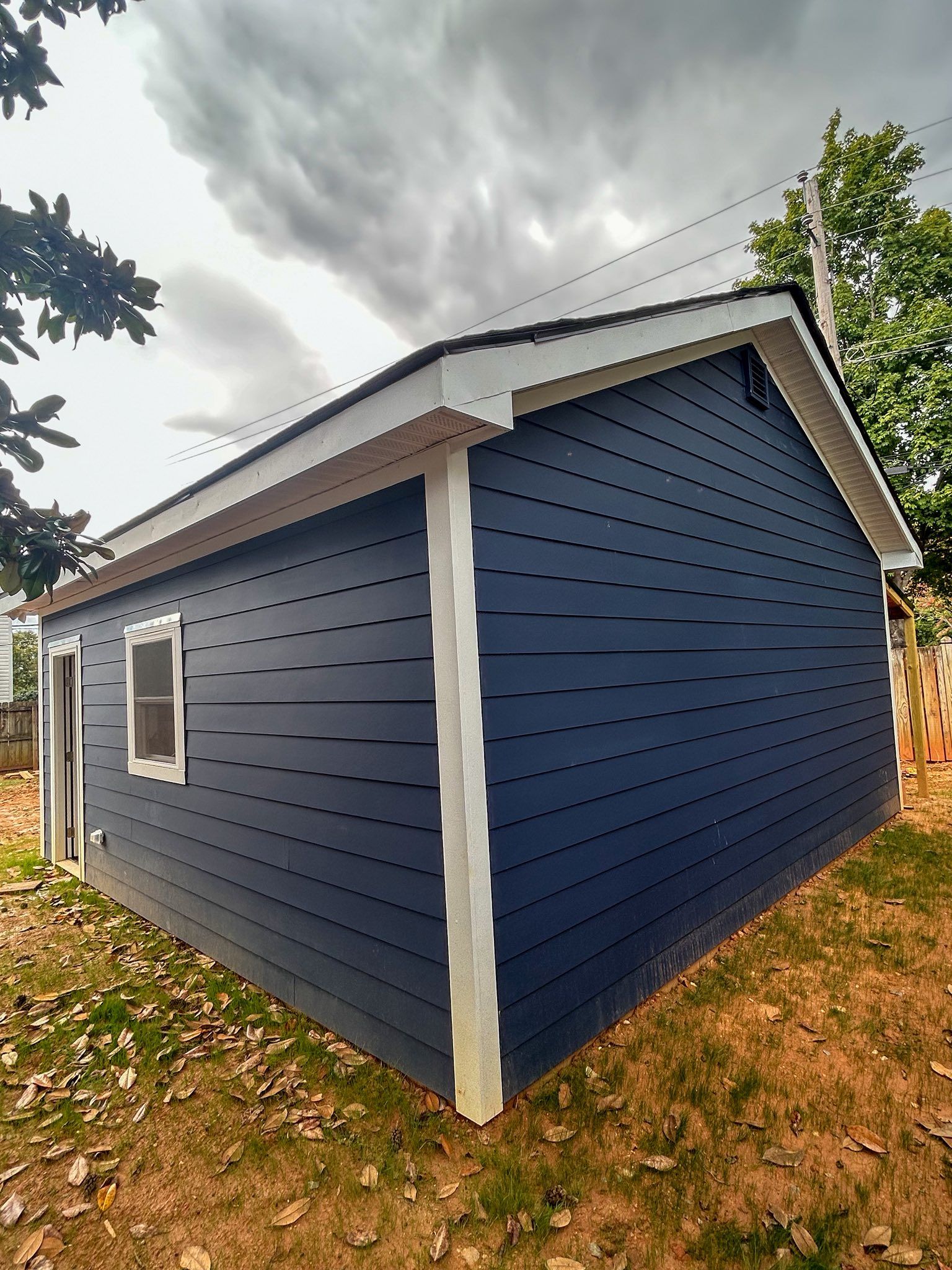A blue shed with white trim and a single window stands on a grassy plot under a cloudy, overcast sky.