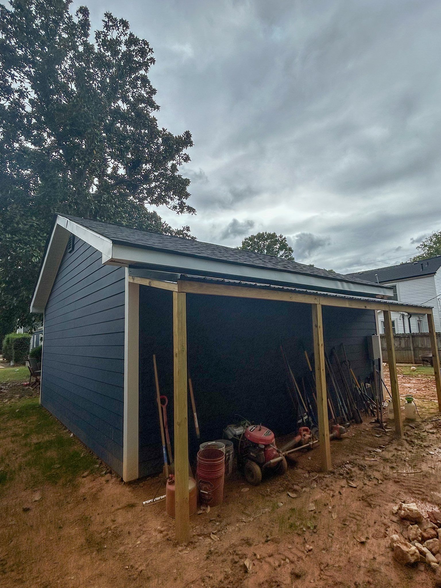 A dark blue shed with a metal roof and a wooden-framed covered side porch area in a yard under a cloudy sky.