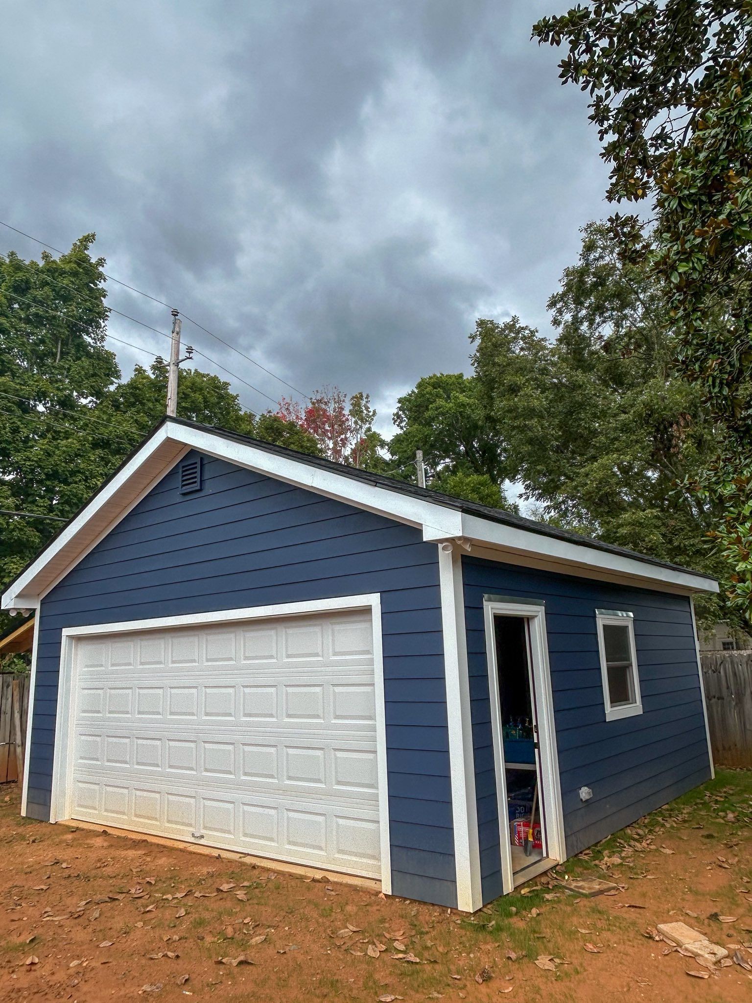 A blue detached garage with white trim, a white garage door, and an open side door, set against trees and a cloudy sky.