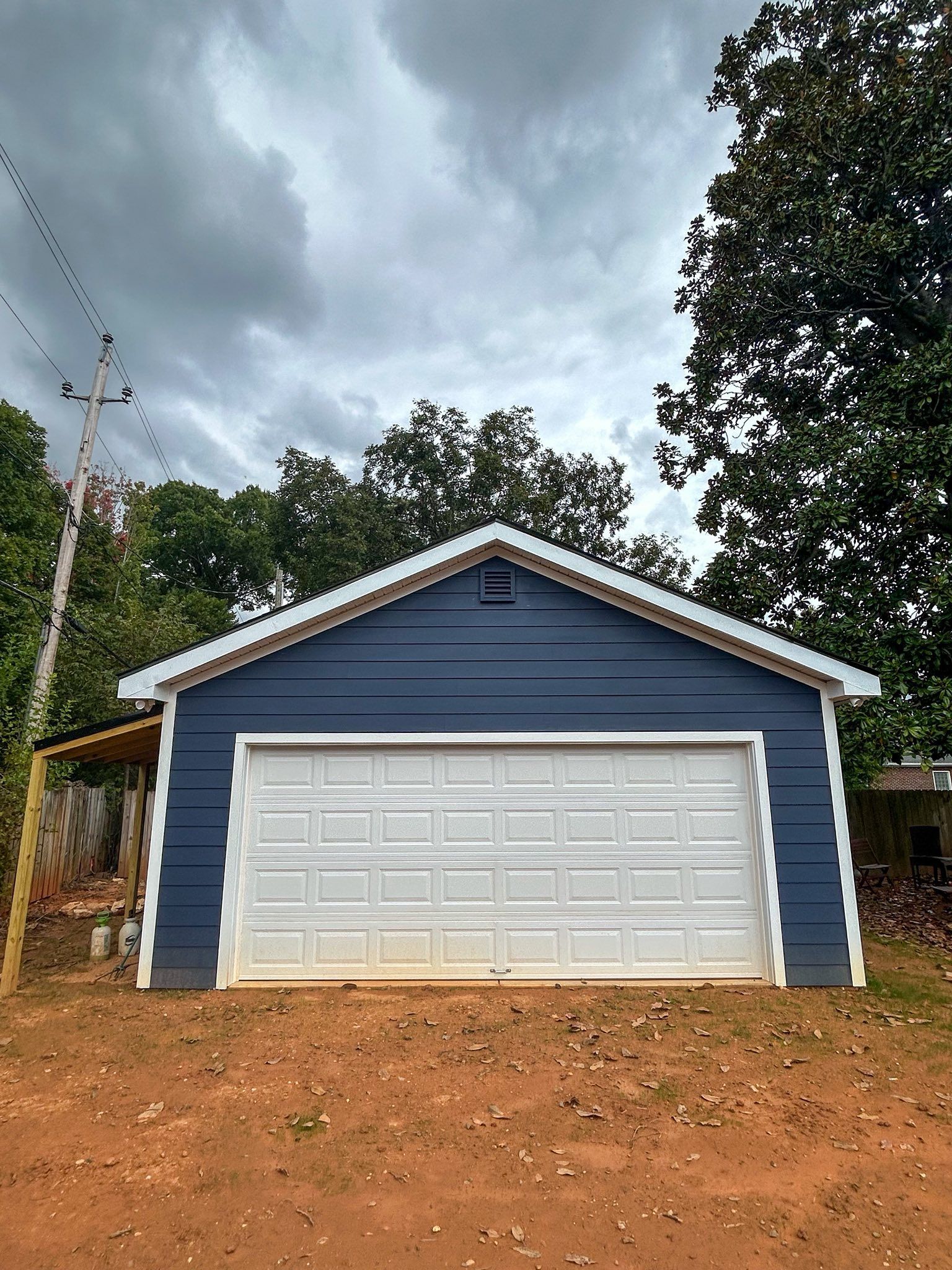A blue, detached garage with a white garage door, white trim, and a gabled roof set against a cloudy sky.