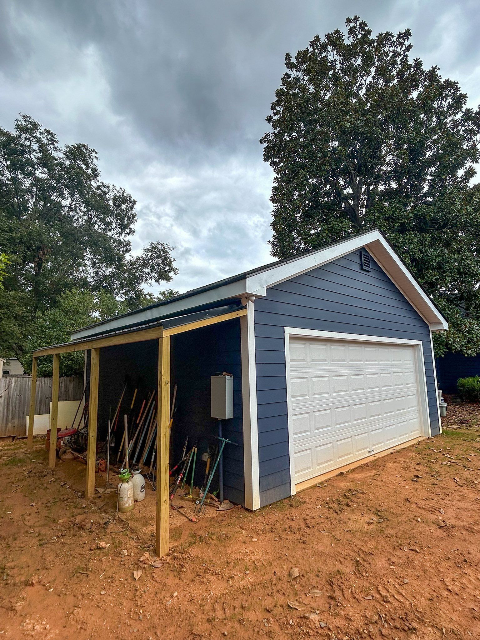 A blue detached garage with a white roll-up door and an attached wooden open-sided shed frame, set on red clay soil.