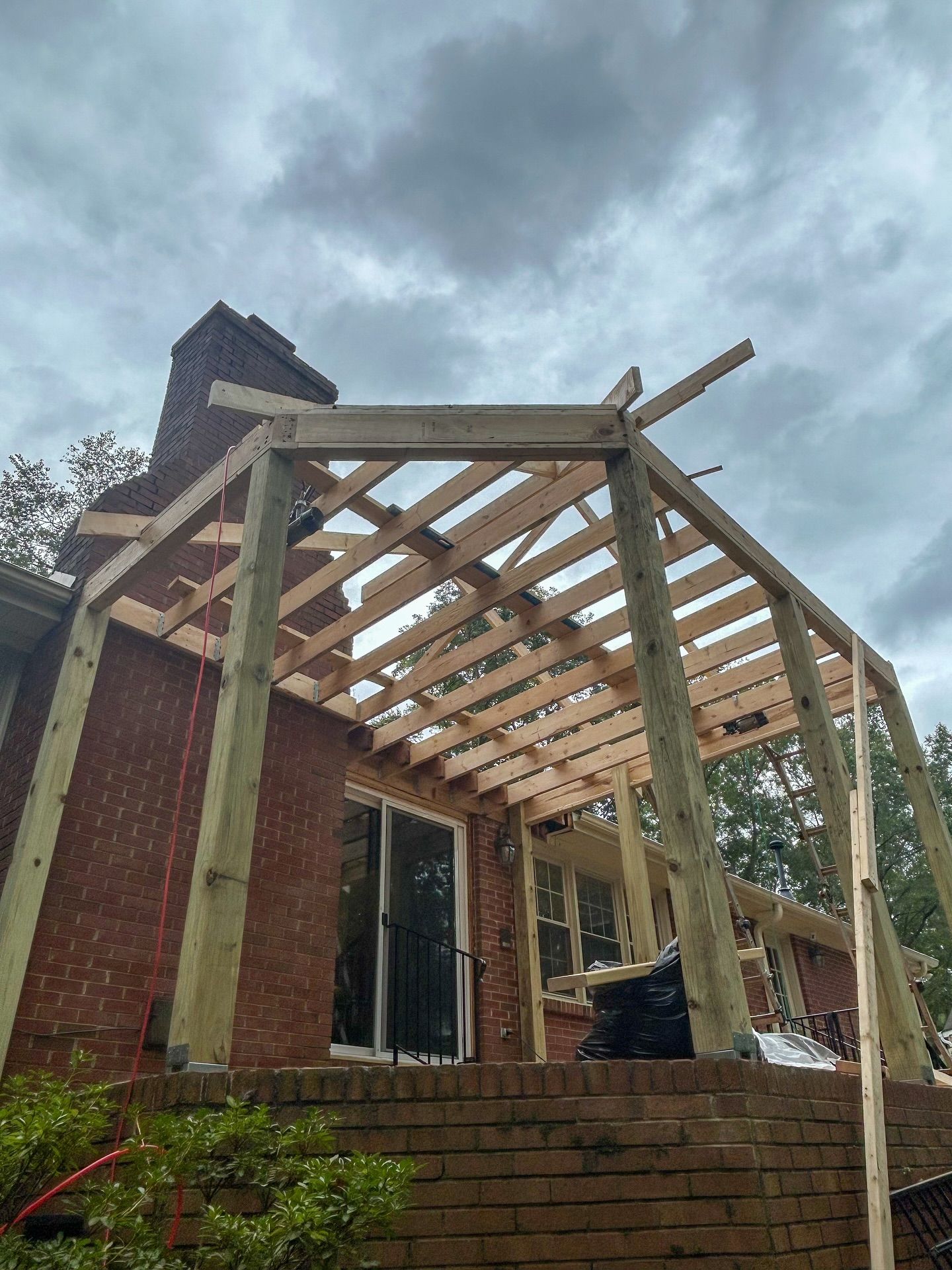 Wooden framing for an open patio cover under construction attached to a red brick house exterior against a cloudy sky.