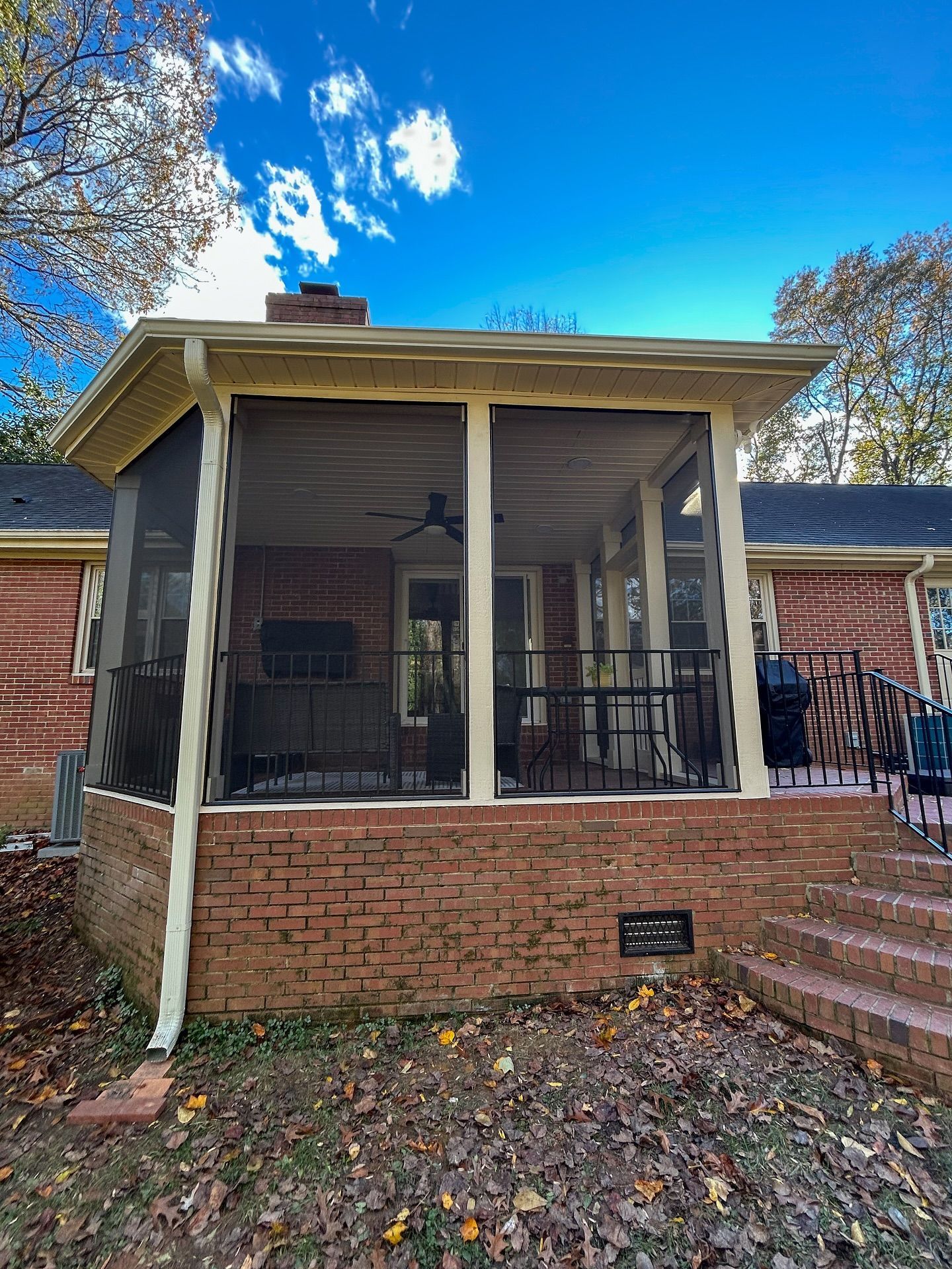 A cream-colored screened-in porch with a brick base and steps, attached to a brick house under a clear blue sky.