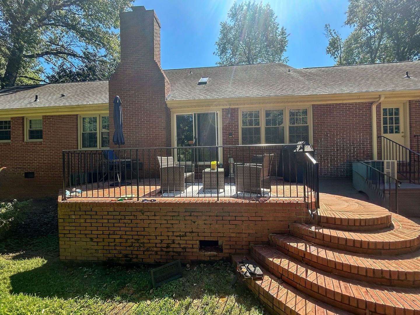 A brick house with a raised brick patio, black metal railings, and curved steps leading to a backyard lawn.