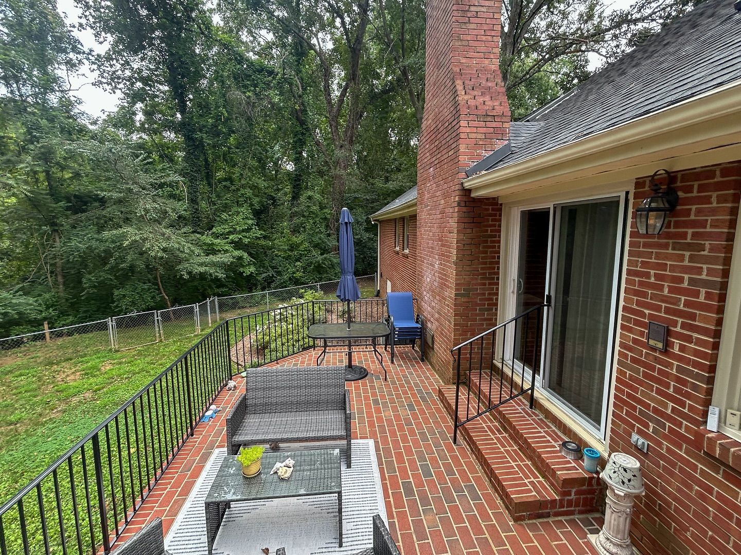 A brick patio with outdoor seating next to a brick house, overlooking a wooded backyard with a black iron railing.