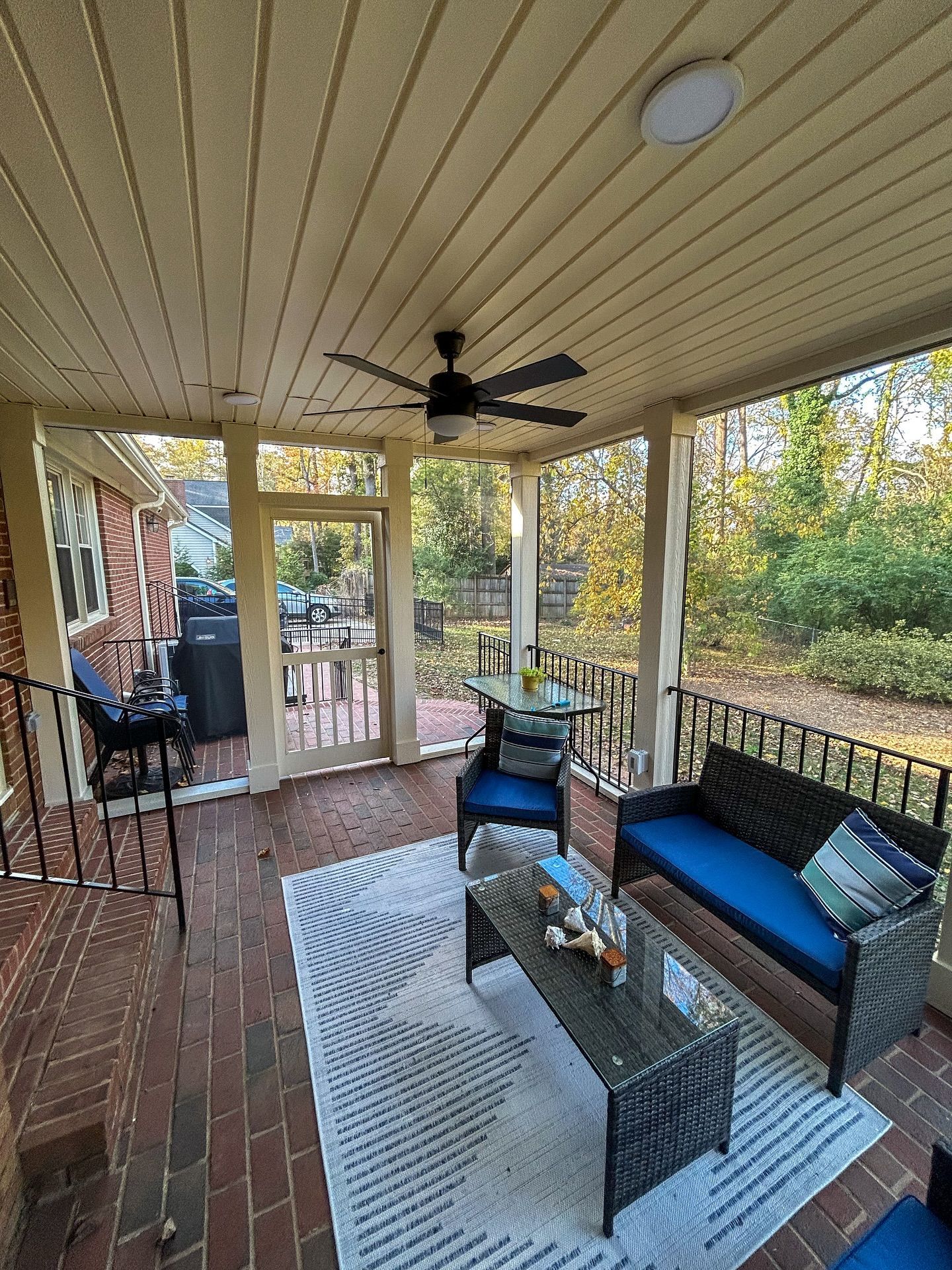 A screened-in porch with brick flooring, a wicker sofa with blue cushions, a coffee table, and a ceiling fan.