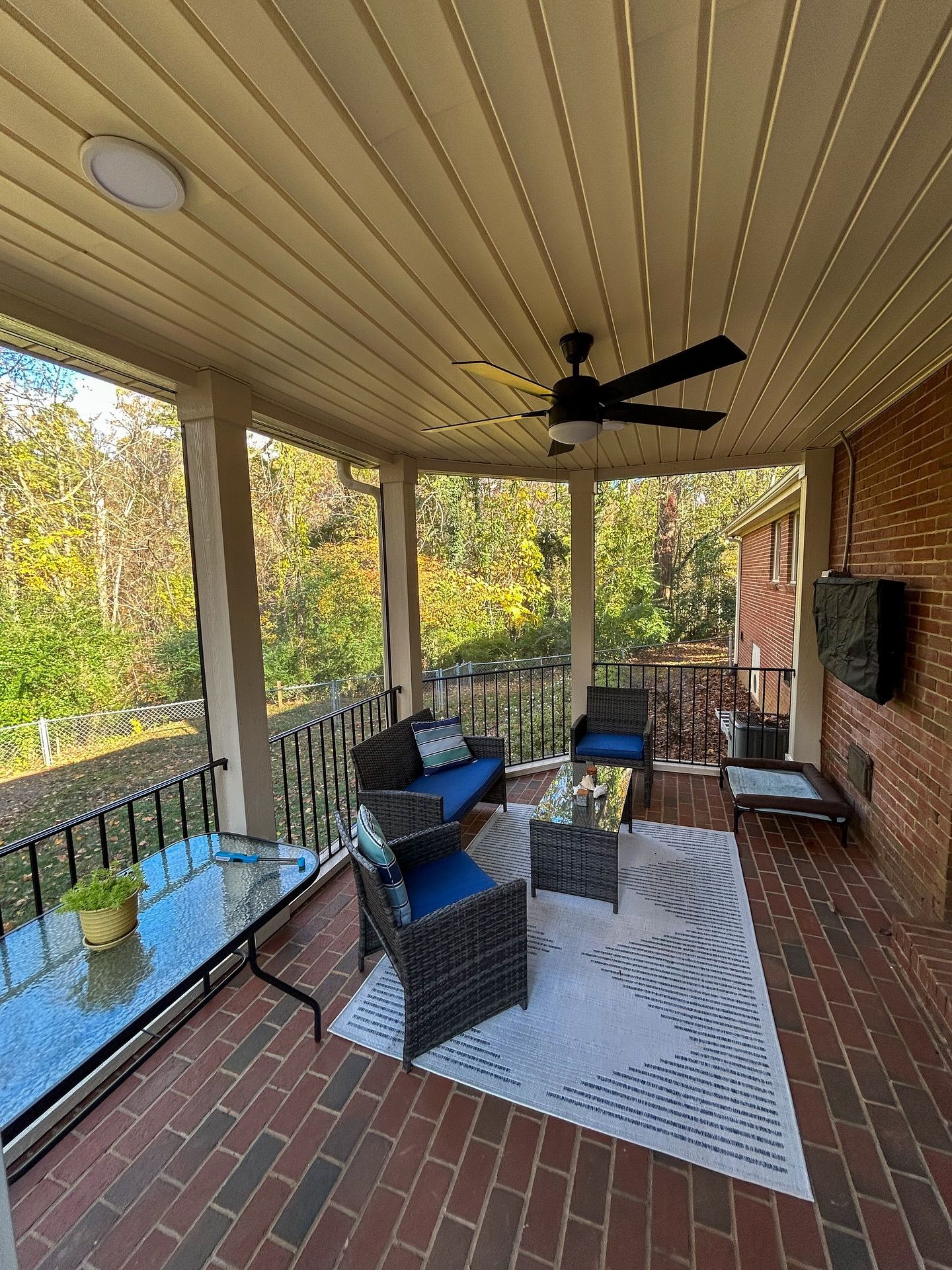 A covered outdoor brick patio with dark wicker seating, a glass-topped table, a white patterned rug, and a ceiling fan.