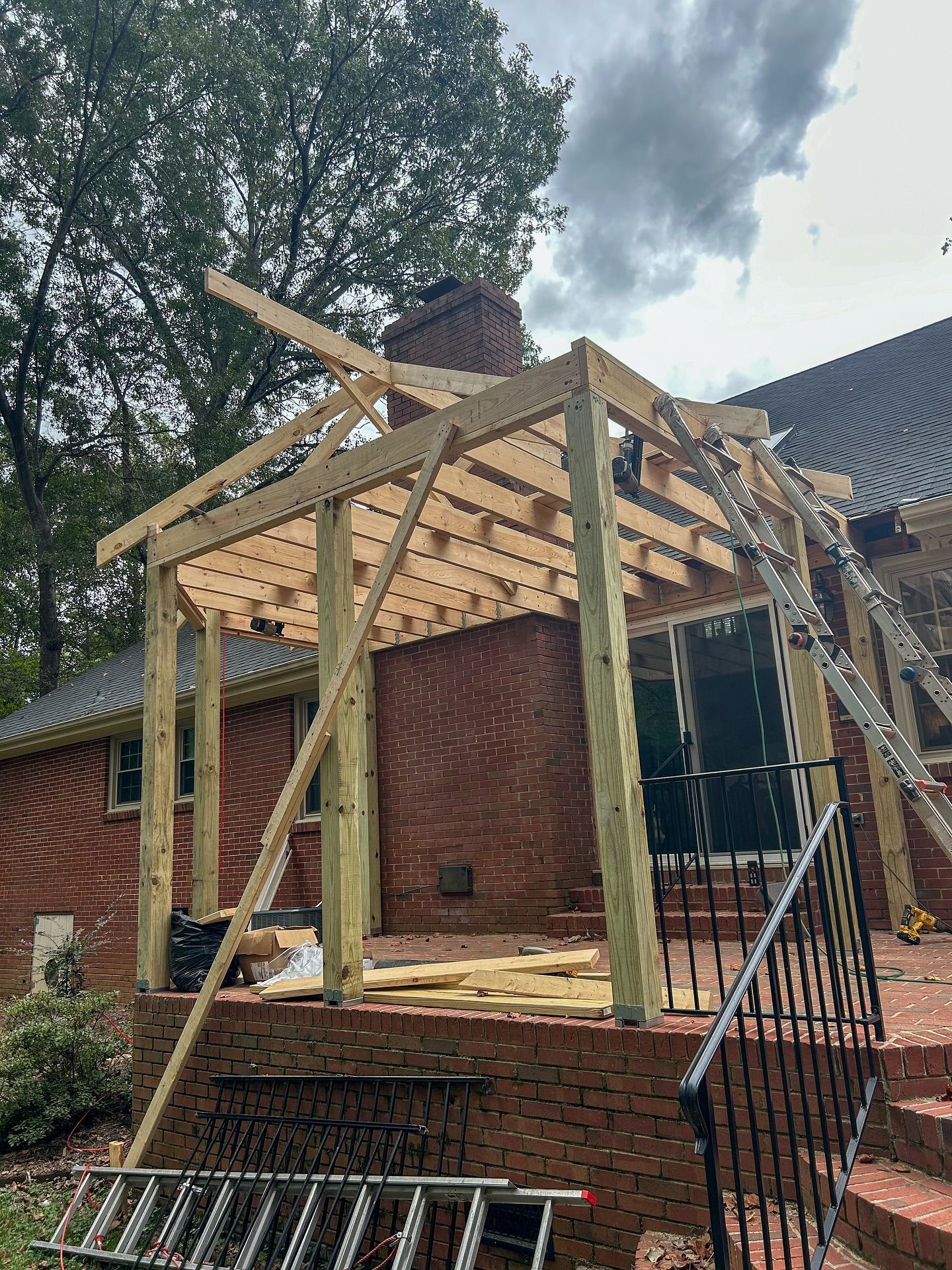 A wooden porch frame under construction against the brick exterior of a house, with a ladder leaning against it.