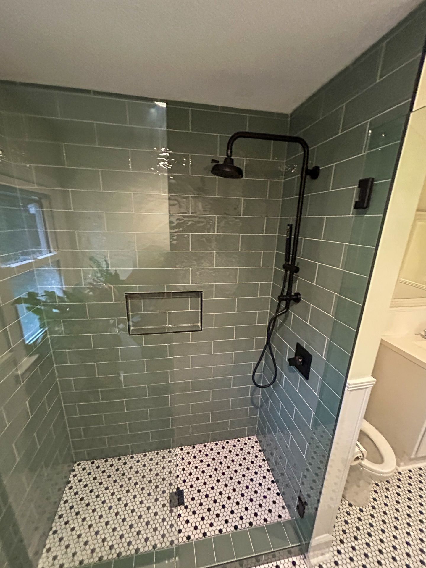 A shower with sage green subway tile walls, a matte black rainfall showerhead, and a black-and-white patterned tile floor.