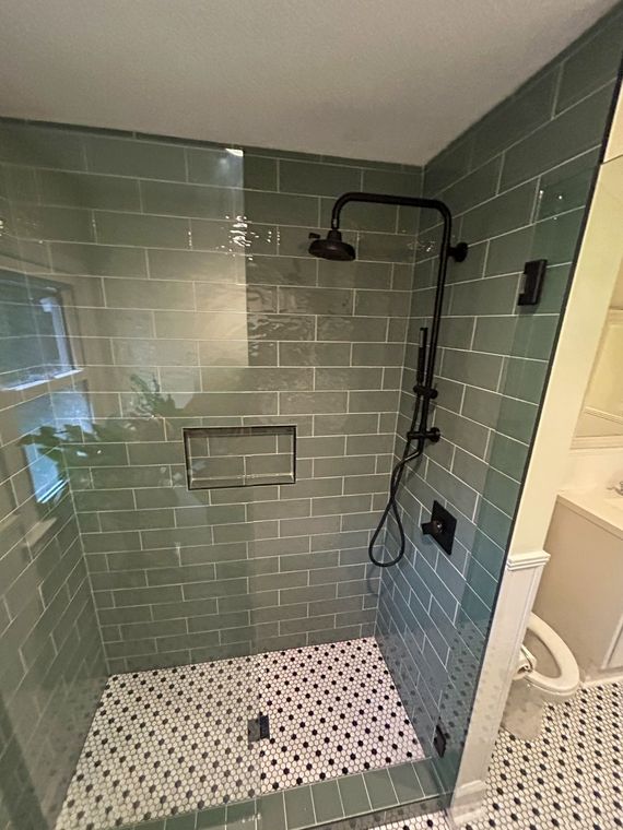 A modern shower with sage green subway tile walls, a matte black shower fixture, and a black-and-white patterned floor.