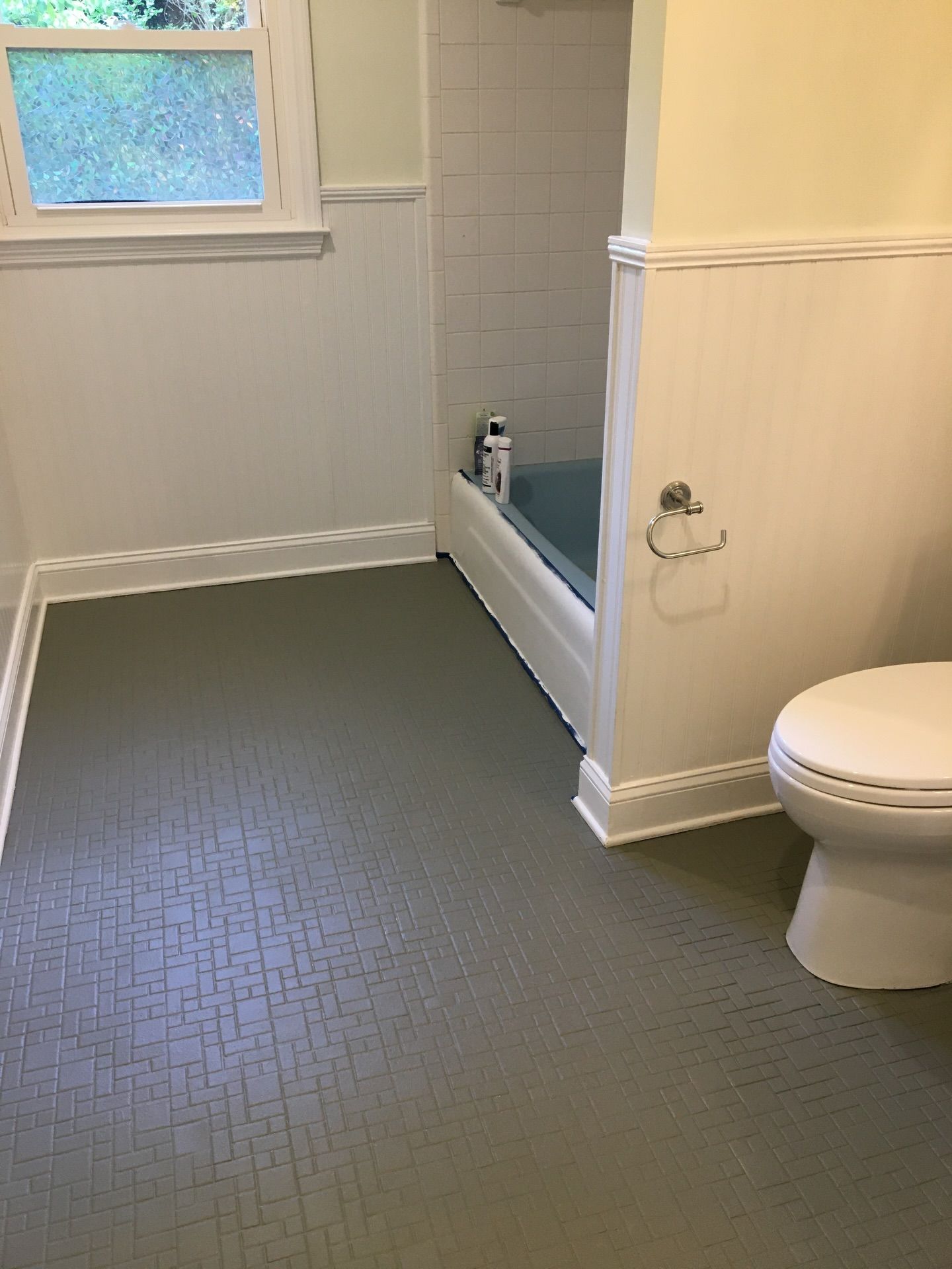 A bathroom with a white toilet, white wainscoting, a blue bathtub, and grey textured flooring by a window.