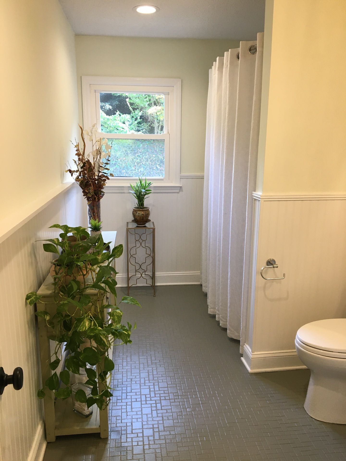 A bathroom interior featuring a grey textured floor, a white shower curtain, a small window, and two potted plants.