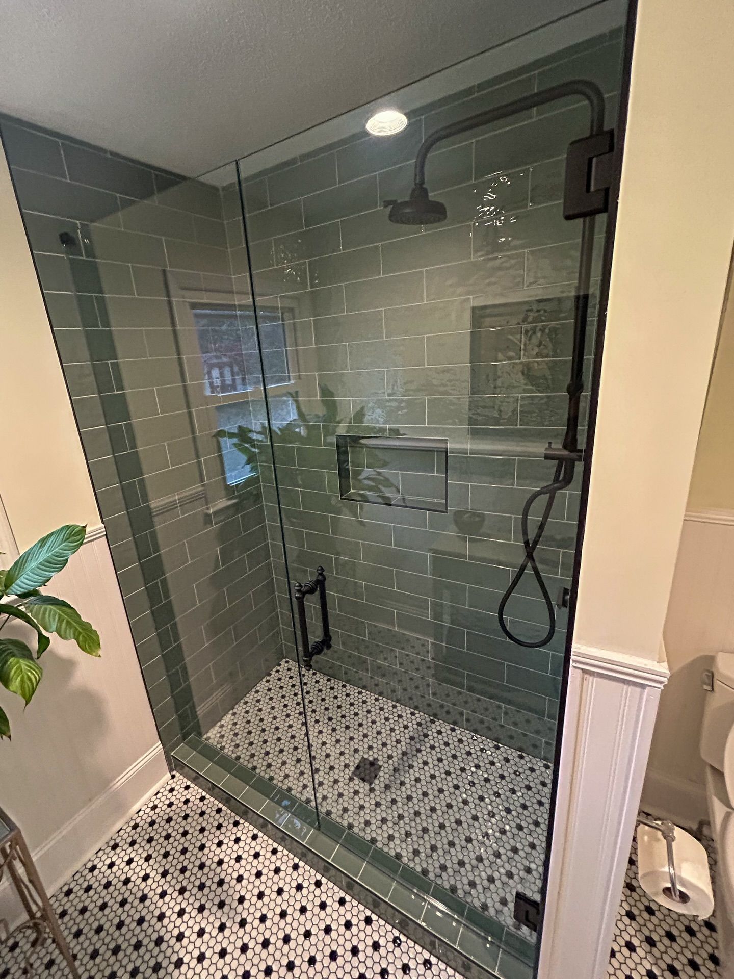 A walk-in shower with gray subway tiles, a black shower head, a glass door, and black-and-white patterned floor tiles.