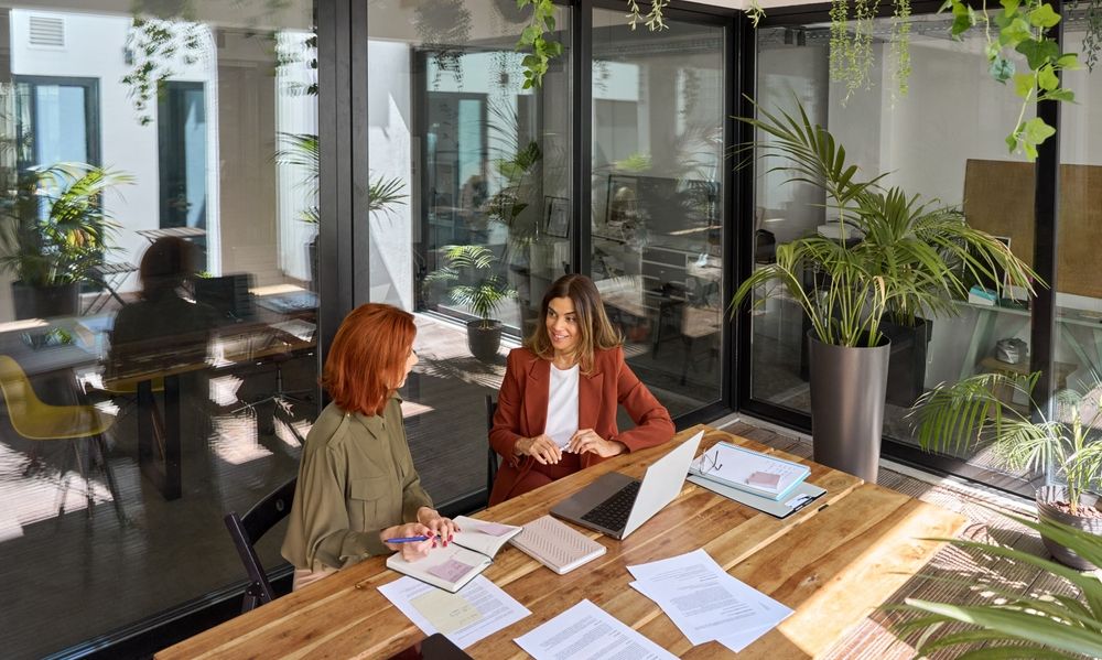 Two women are sitting at a table in an office with a laptop.