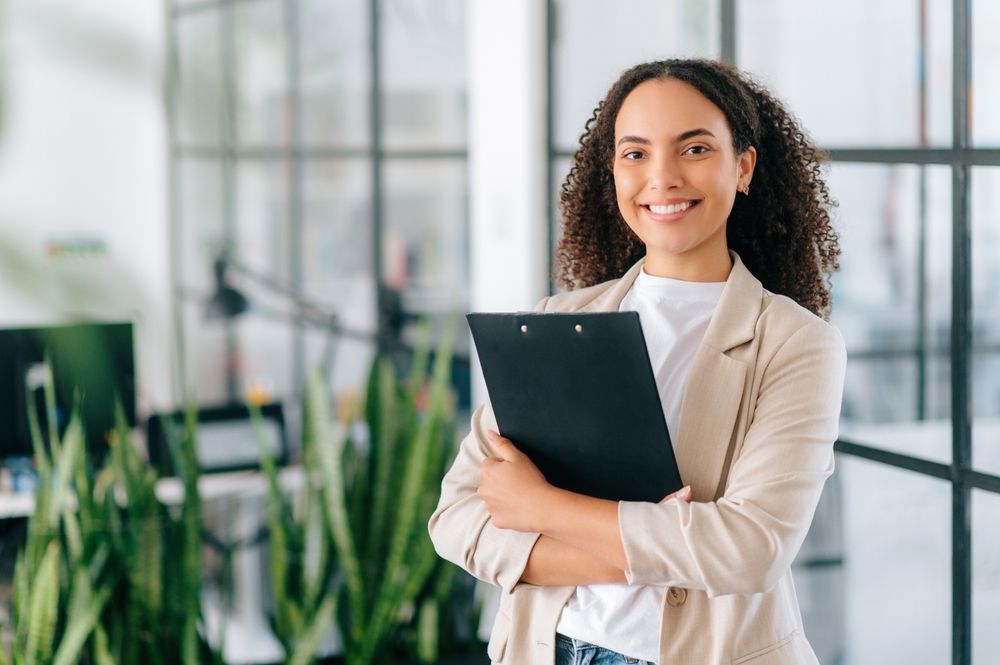 A woman is holding a clipboard and smiling in an office.