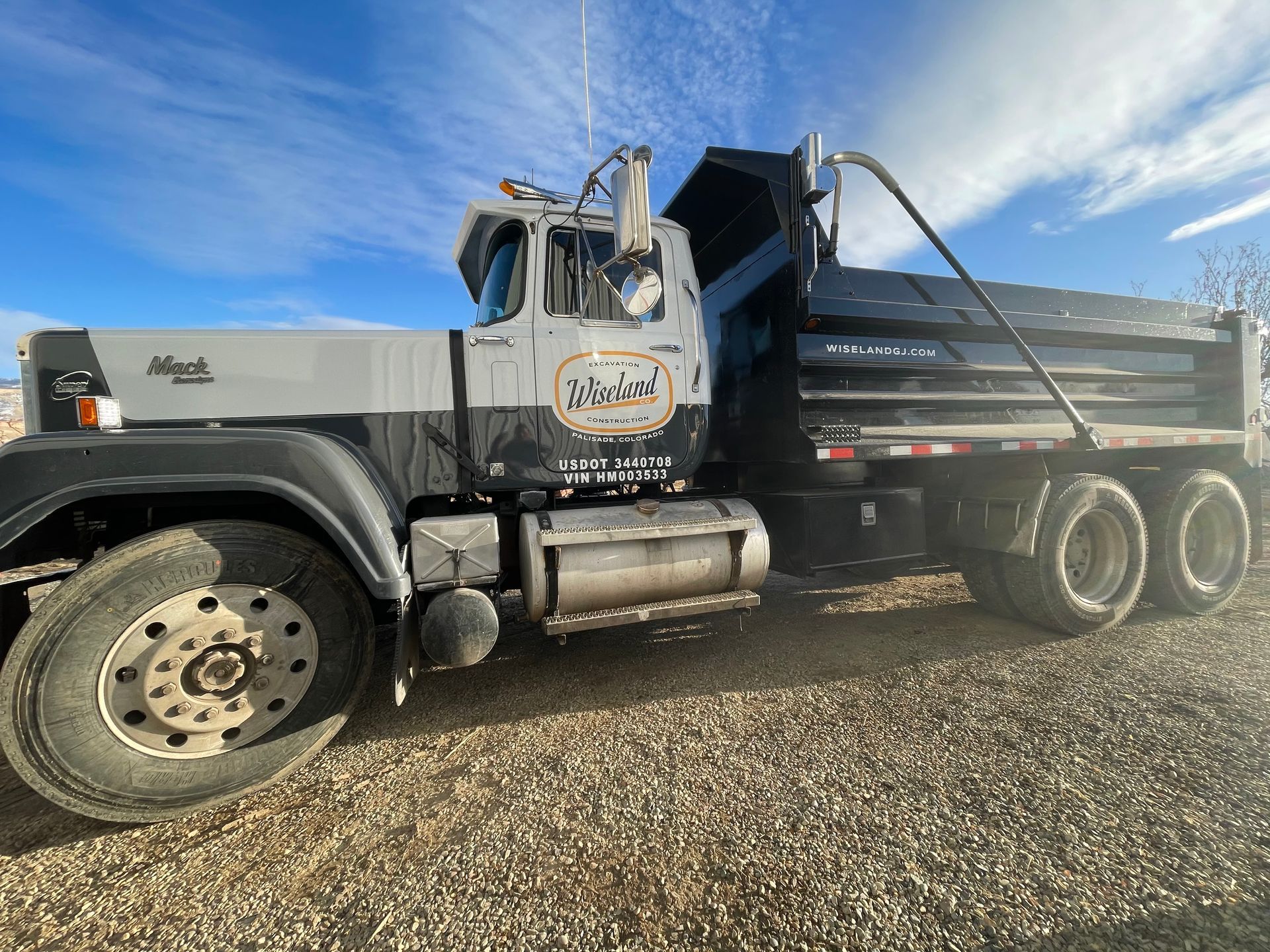 Gray Mack dump truck parked on gravel with a raised bed under a blue sky.