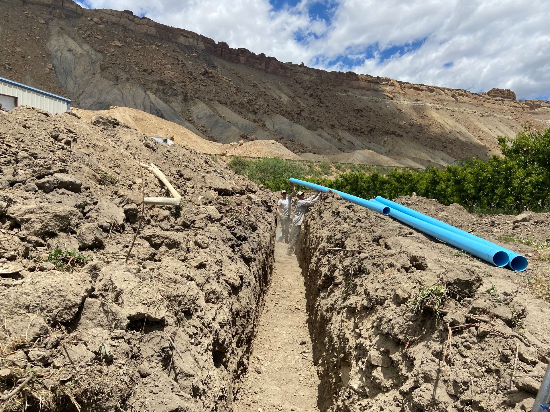 A man is standing in a trench with blue pipes in the dirt.
