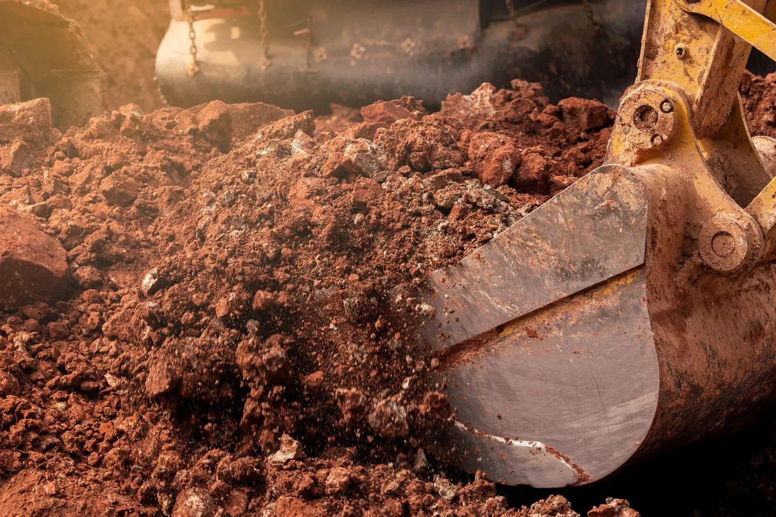 Excavator bucket filled with reddish-brown soil, partially obscuring the machine in a construction site.