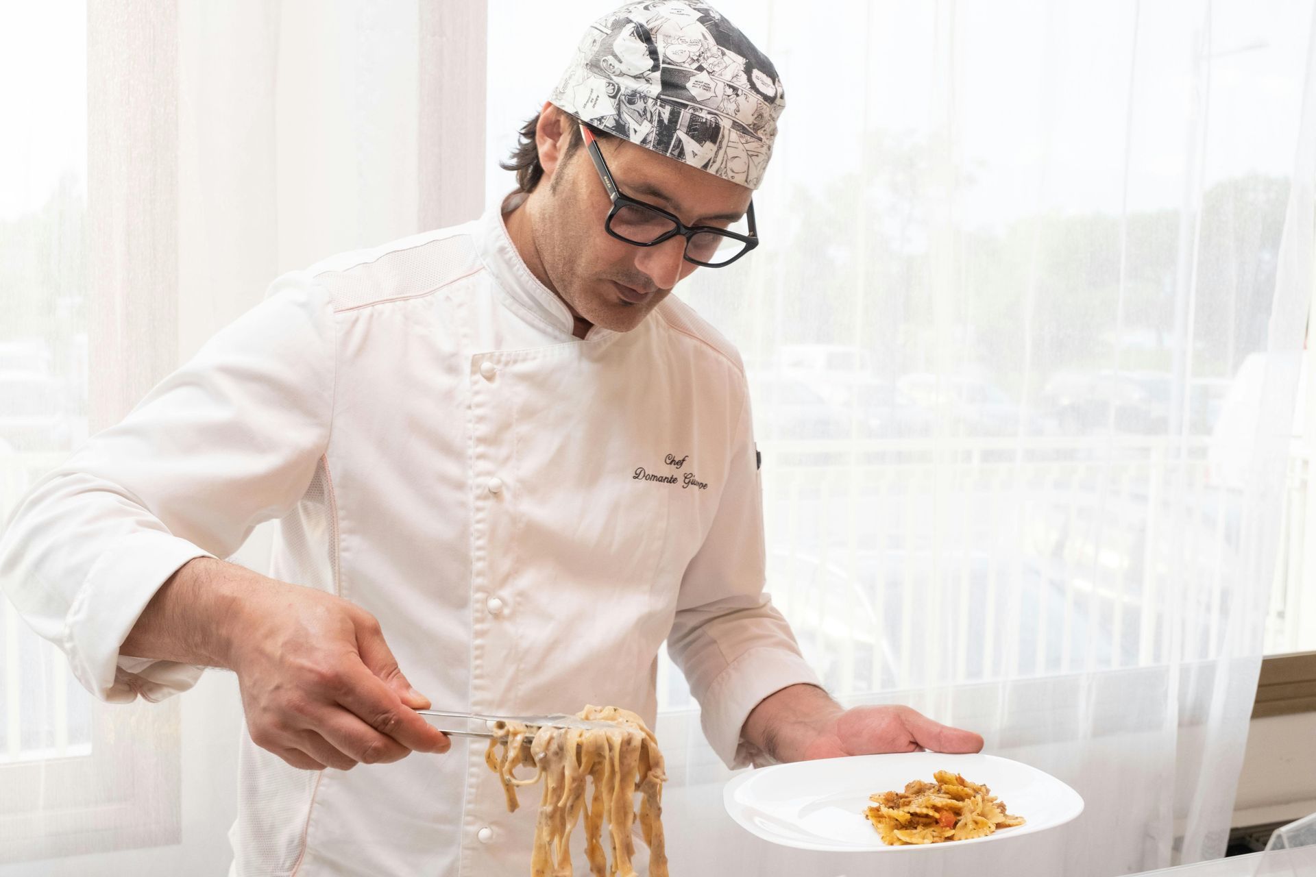 Lo chef impiatta i noodles in una cucina luminosa, indossando un'uniforme bianca e un cappello decorato.