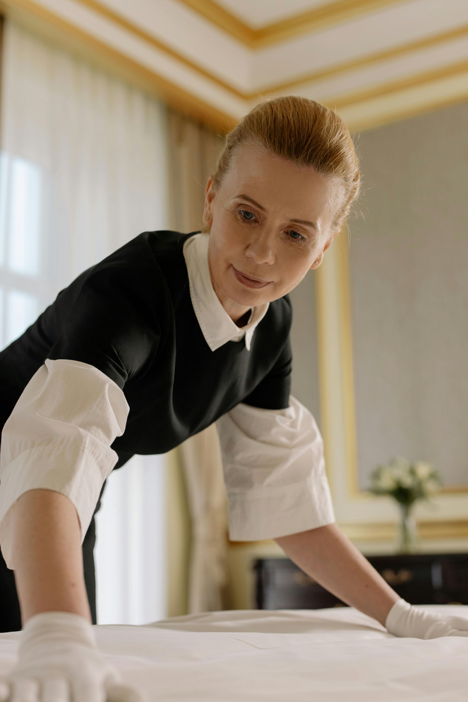 Una cameriera d'albergo in uniforme bianca e nera rifà un letto in una stanza luminosa.