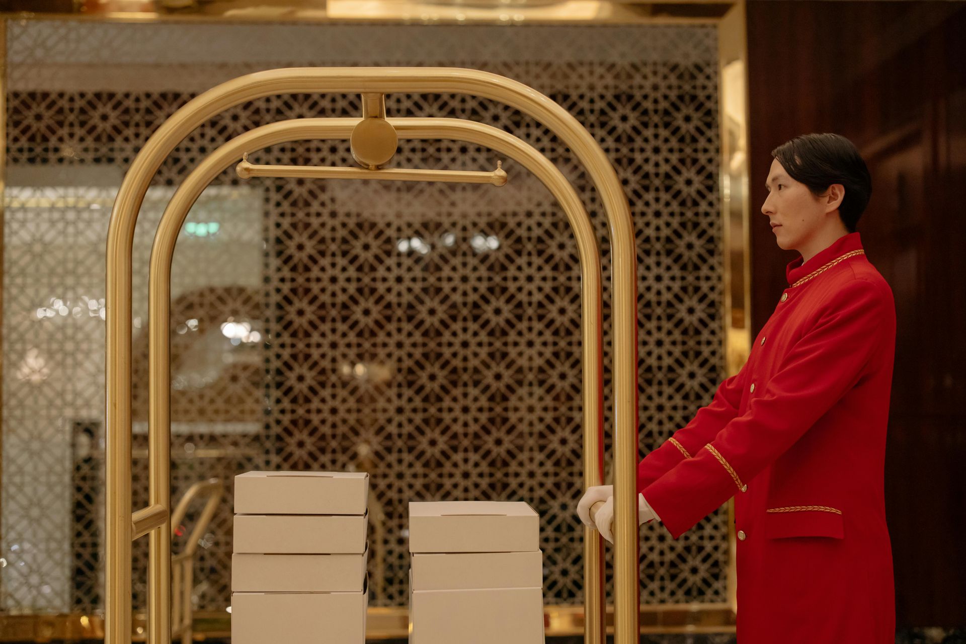Una donna con un cappotto rosso spinge un carrello portabagagli dorato nella hall decorata di un hotel.