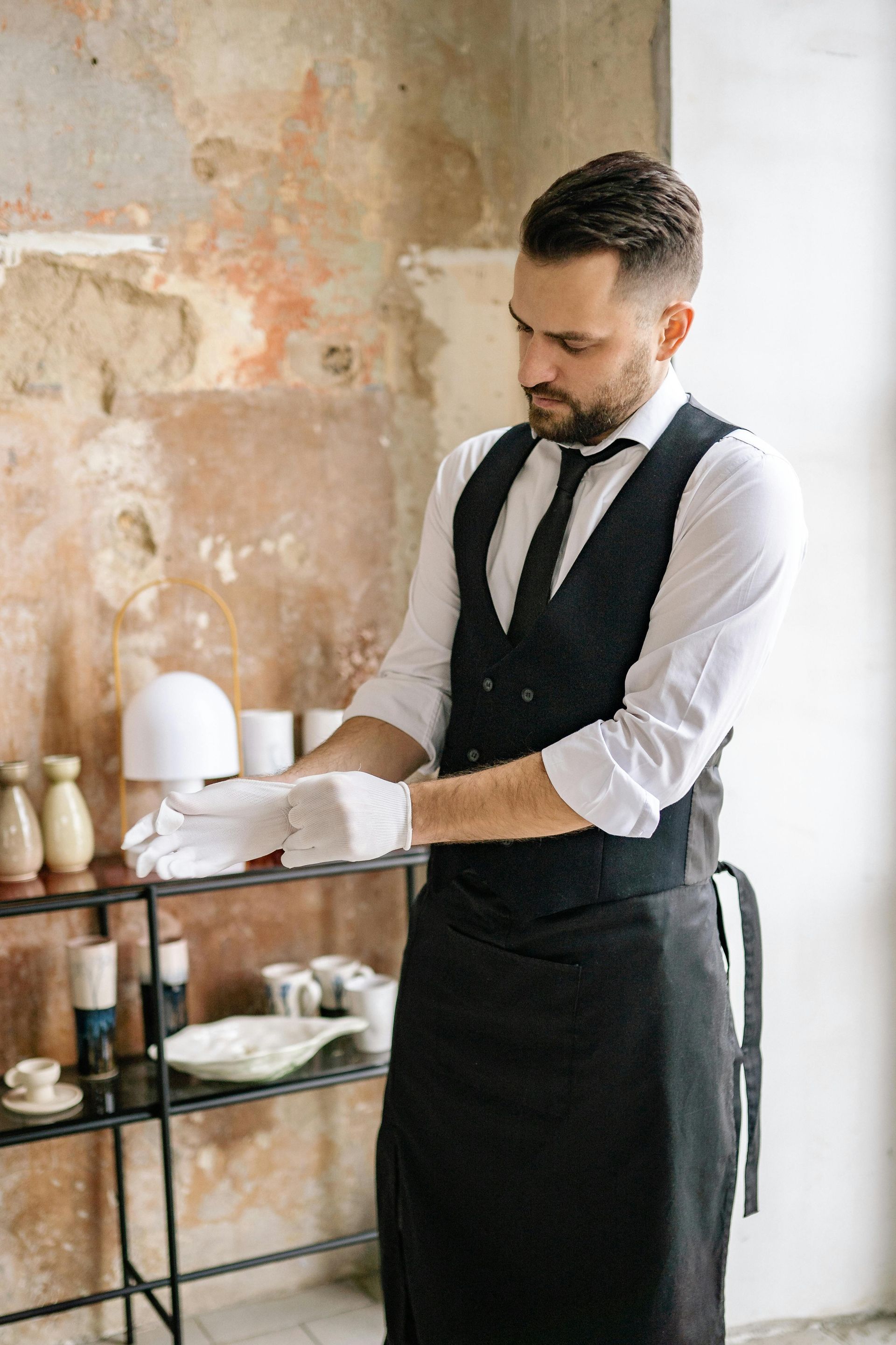 Cameriere con gilet nero che sistema le posate a un tavolino di un caffè accanto a una parete con una texture particolare.
