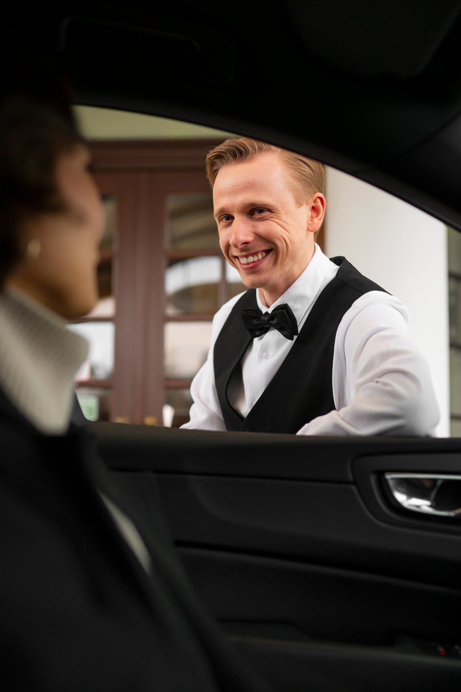 Man in formal wear smiles, leaning into a car window, interacting with a person inside.