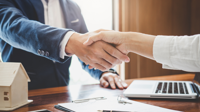 Two people shaking hands over a desk; miniature house and laptop in view.