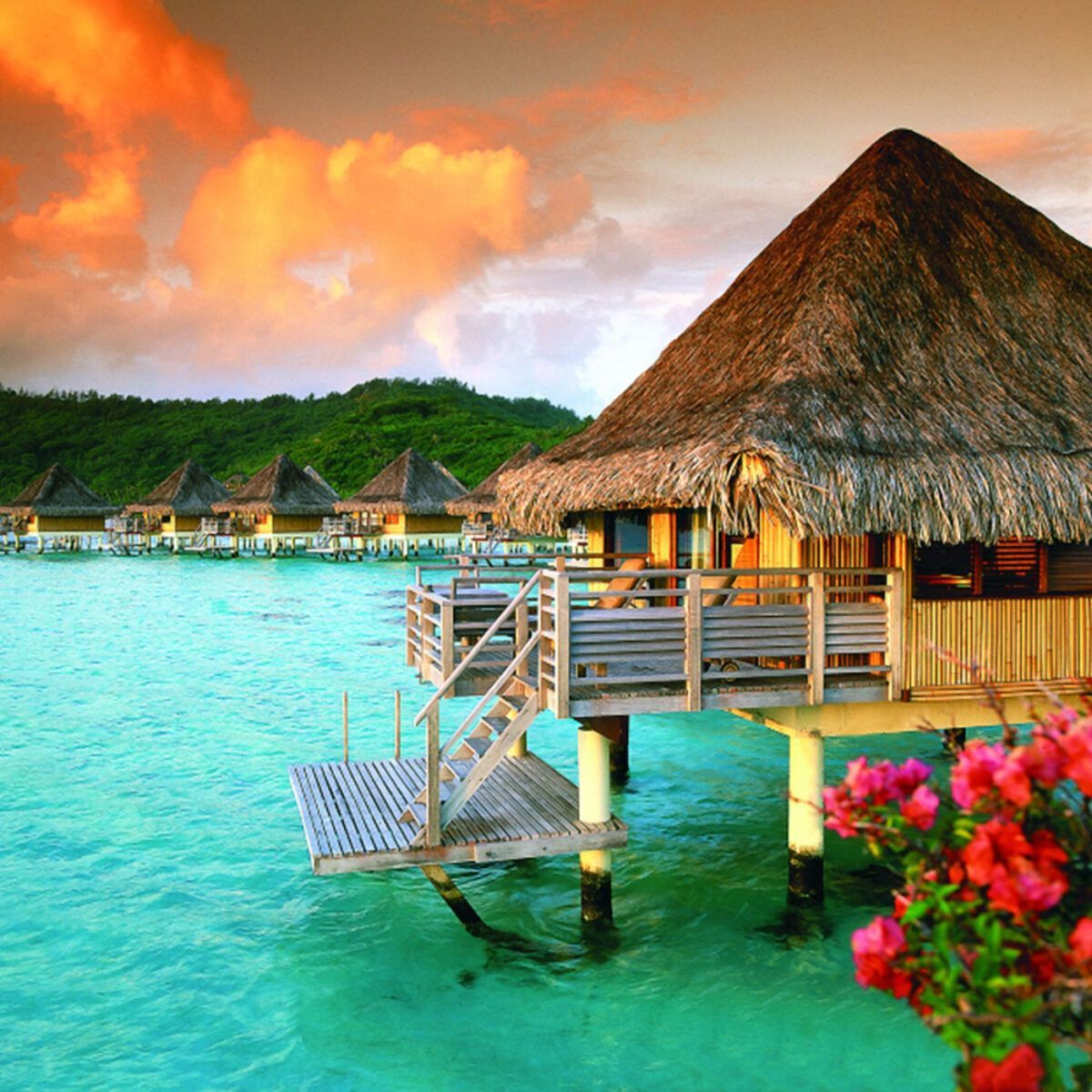 Overwater bungalow with thatched roof, turquoise water, pink flowers, and sunset sky.