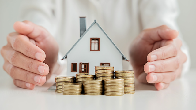 Hands protect a miniature house and stacks of gold coins on a white surface.