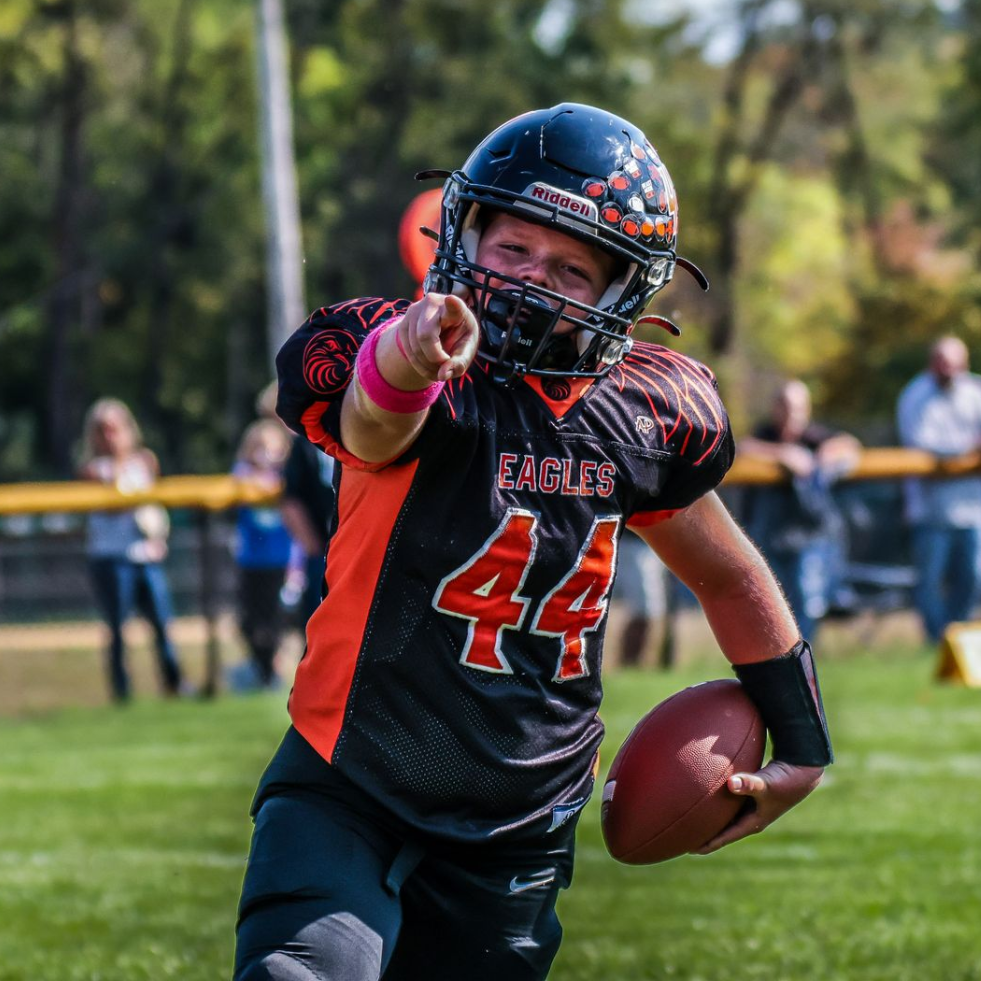 Football players in action on a grassy field; one runs with the ball, pursued by two opponents.