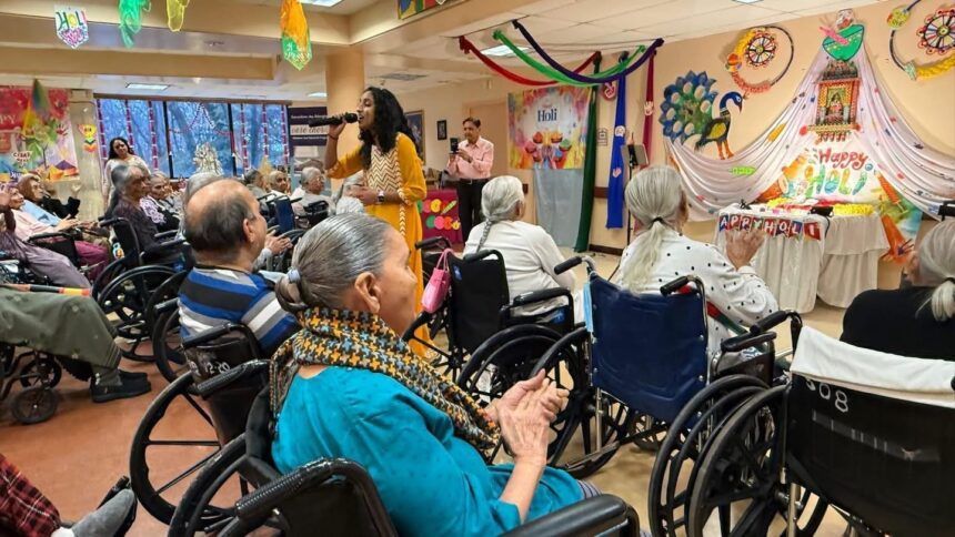 A woman performs with a microphone for a seated audience in wheelchairs at a festive, decorated indoor event.