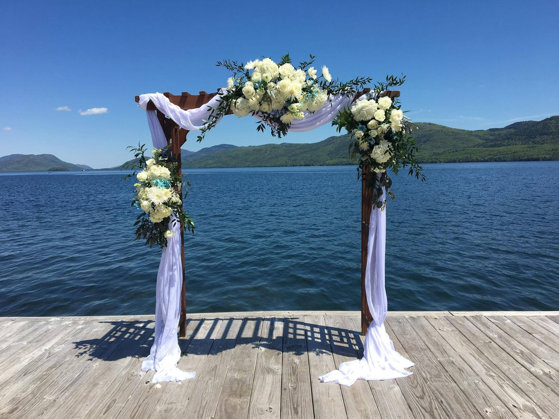 a wooden arch decorated with white flowers is on a dock overlooking a lake .
