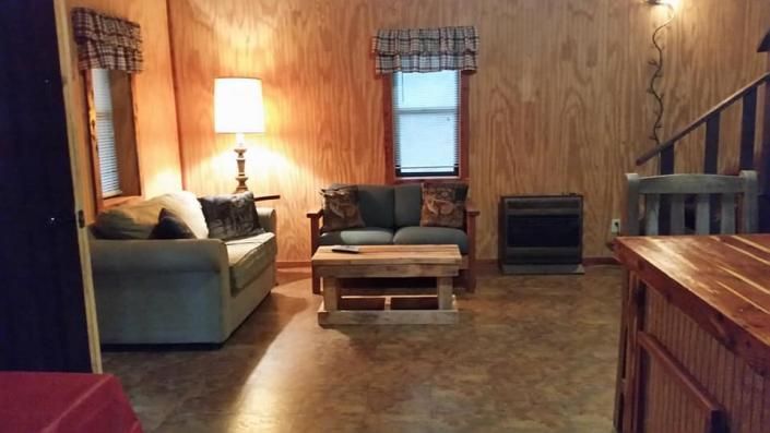 Cozy cabin-style living room with sofa, armchairs, coffee table, lamp, and wood-paneled walls