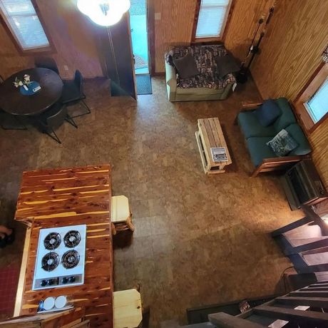 Cozy cabin living room with wood-paneled walls, sofa, armchairs, small table, and kitchen island viewed from above