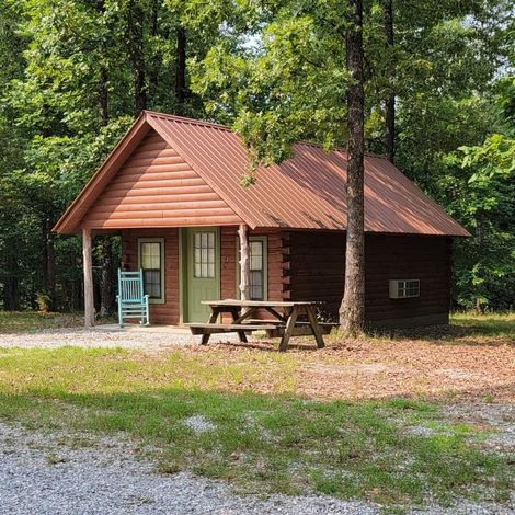 Small wooden cabin with a red roof in a wooded clearing, with a picnic table in front.