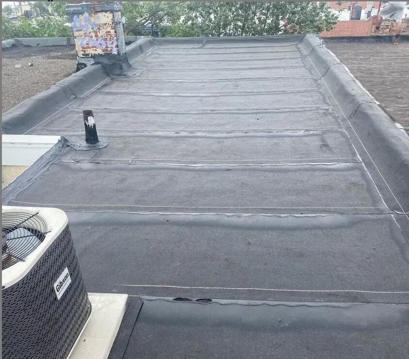 A flat, dark gray roof with horizontal seams, a vent pipe, and an air conditioning unit.