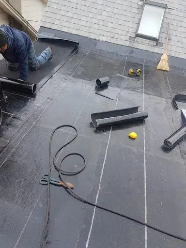 Roofer installing black roofing material on a gray shingle roof. Tools and a skylight are present.