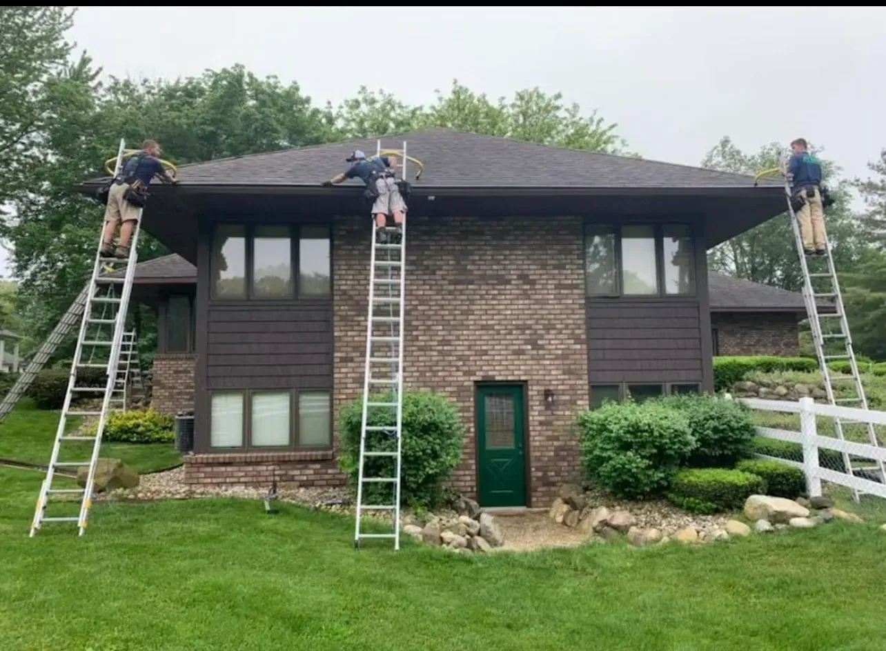 Three people on ladders cleaning gutters on a brown brick house with a green door; cloudy sky.