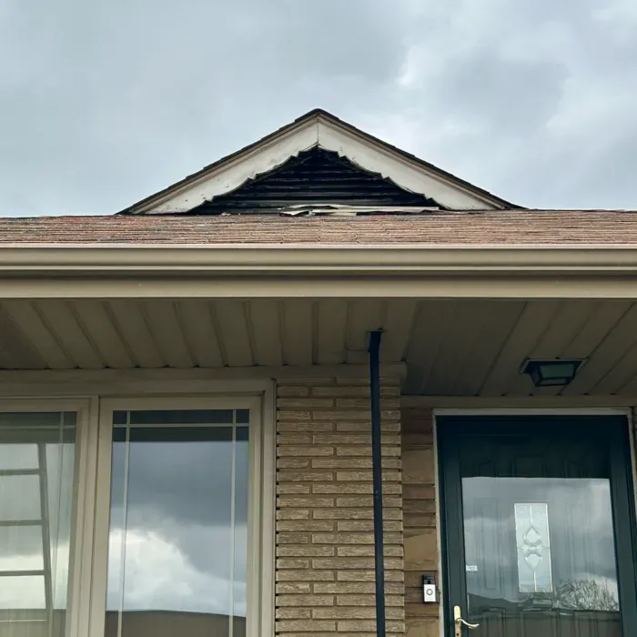 Front of a brick house with damaged roof. Brown roof, beige gutter, a dark front door, and a cloudy sky.