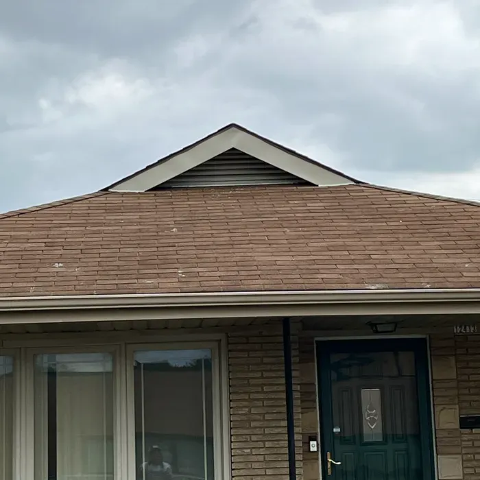 Close-up of a house exterior with a brown roof and a triangle vent beneath a cloudy sky.