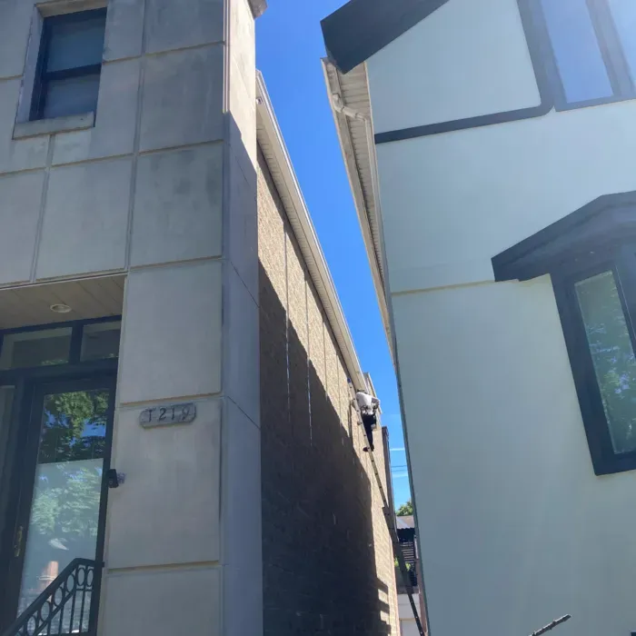 Person on ladder cleaning gutter between two buildings. Sunny day, blue sky.