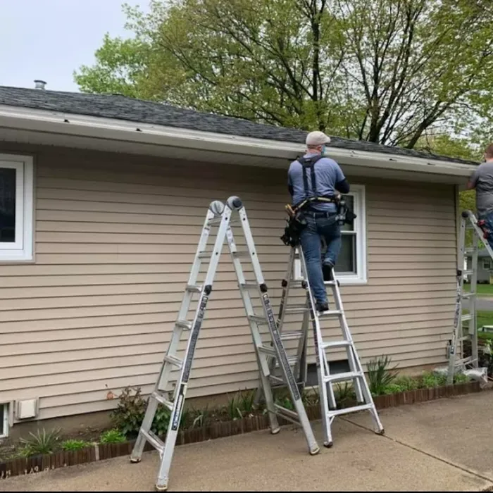 Two people on ladders cleaning gutters of a light brown house with a white trim.