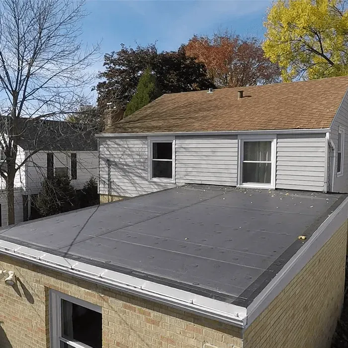 Flat black roof on a brick building, attached to a house with a brown shingled roof, trees in the background.